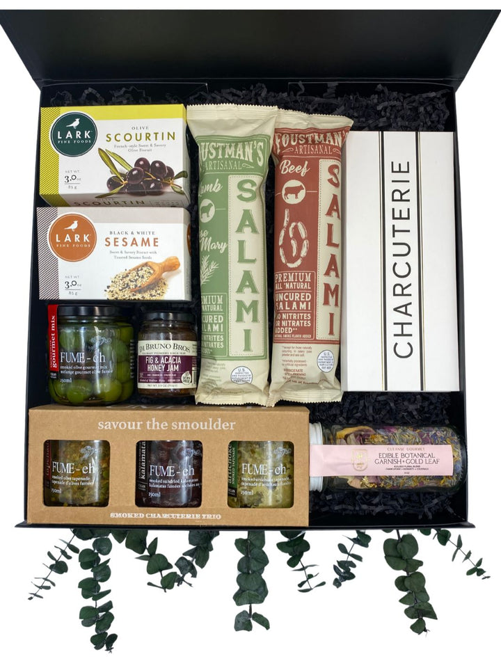 Charcuterie Essentials Gift Box packed with list pad, tapenade trio, smoked olives, fig and honey jam, savory biscuits, salami, and edible botanical garnish.