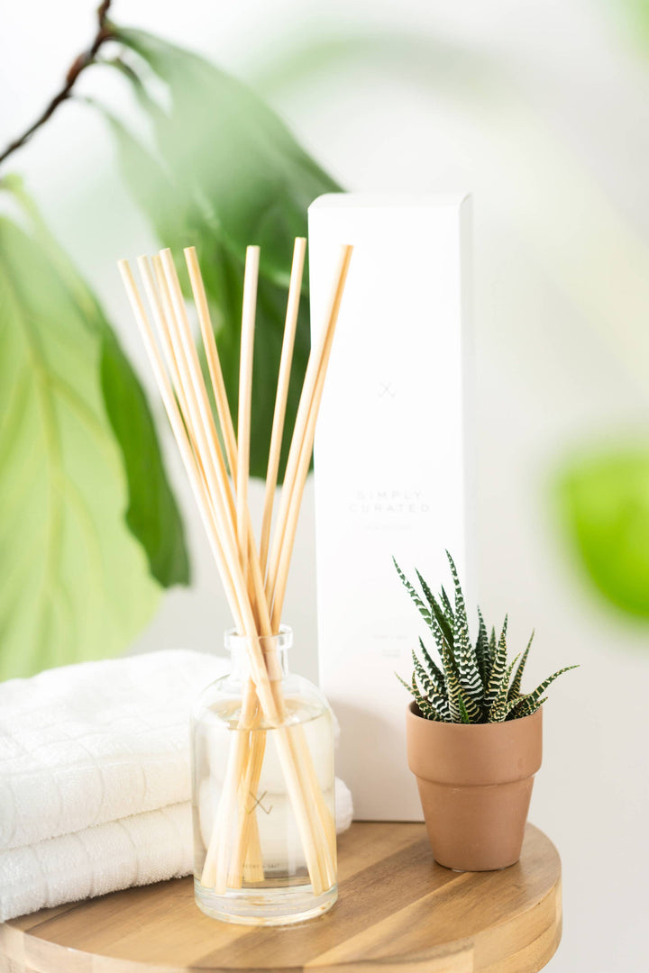 Guava Fig Reed Diffuser on a wooden table, accompanied by soft white towels and a green potted cactus, enhancing a modern home décor aesthetic.