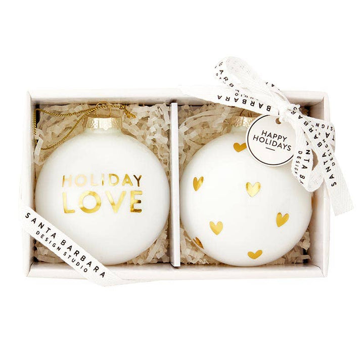 Two glass ornaments, one with “Holiday Love” in gold script and one decorated with gold hearts, presented in a gift box.